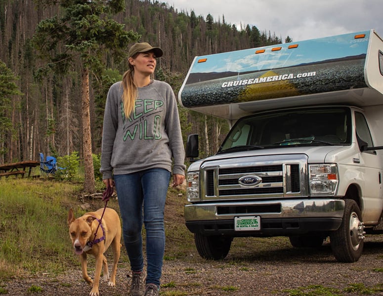 A woman and a dog on a walk near their Cruise America RV.