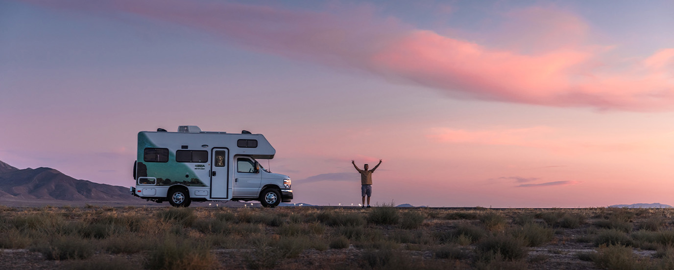 RV parked on road in ping sunset with man standing in front with arms raised